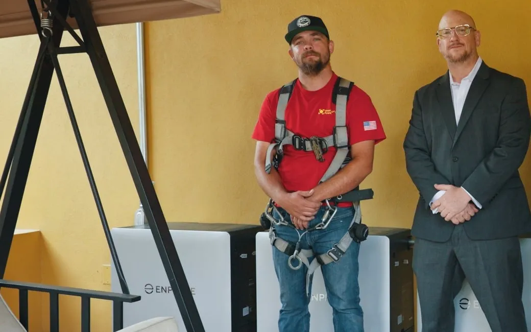 A Westhaven installer and an energy consultant stand beside white Enphase battery units mounted on a yellow patio wall.